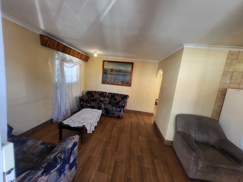 A living room featuring wood-look tiled flooring, cove cornicing, and a timber window pelmet, presenting a dated aesthetic in need of cosmetic updates.