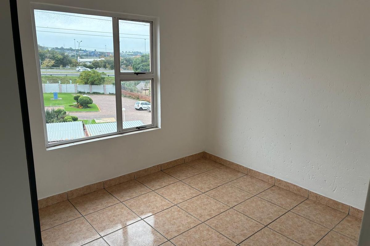 A bright, empty bedroom featuring neutral tiled flooring and a large aluminium-framed window with views of the surrounding complex.