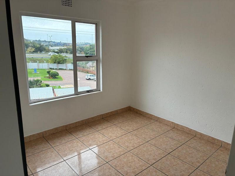 A bright, empty bedroom featuring neutral tiled flooring and a large aluminium-framed window with views of the surrounding complex.