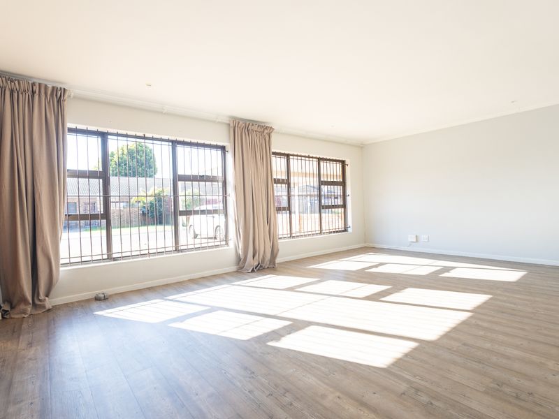A bright, spacious, and empty living room featuring light-toned laminate flooring and large windows fitted with security bars.