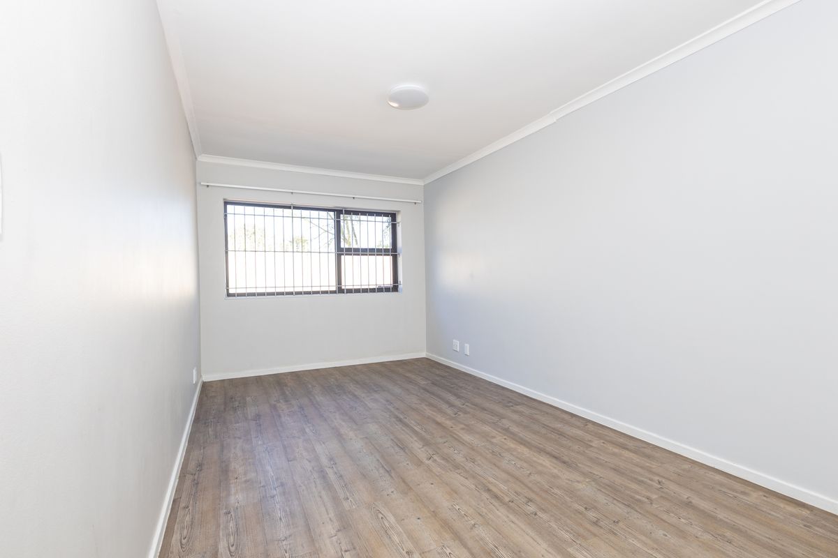 A bright, empty bedroom featuring modern laminate flooring, neutral walls, and a window fitted with security bars.