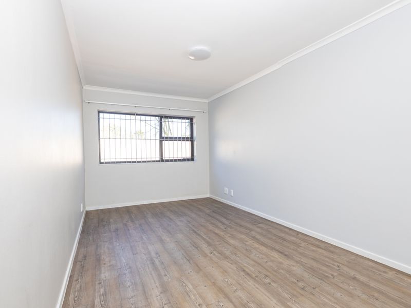 A bright, empty bedroom featuring modern laminate flooring, neutral walls, and a window fitted with security bars.