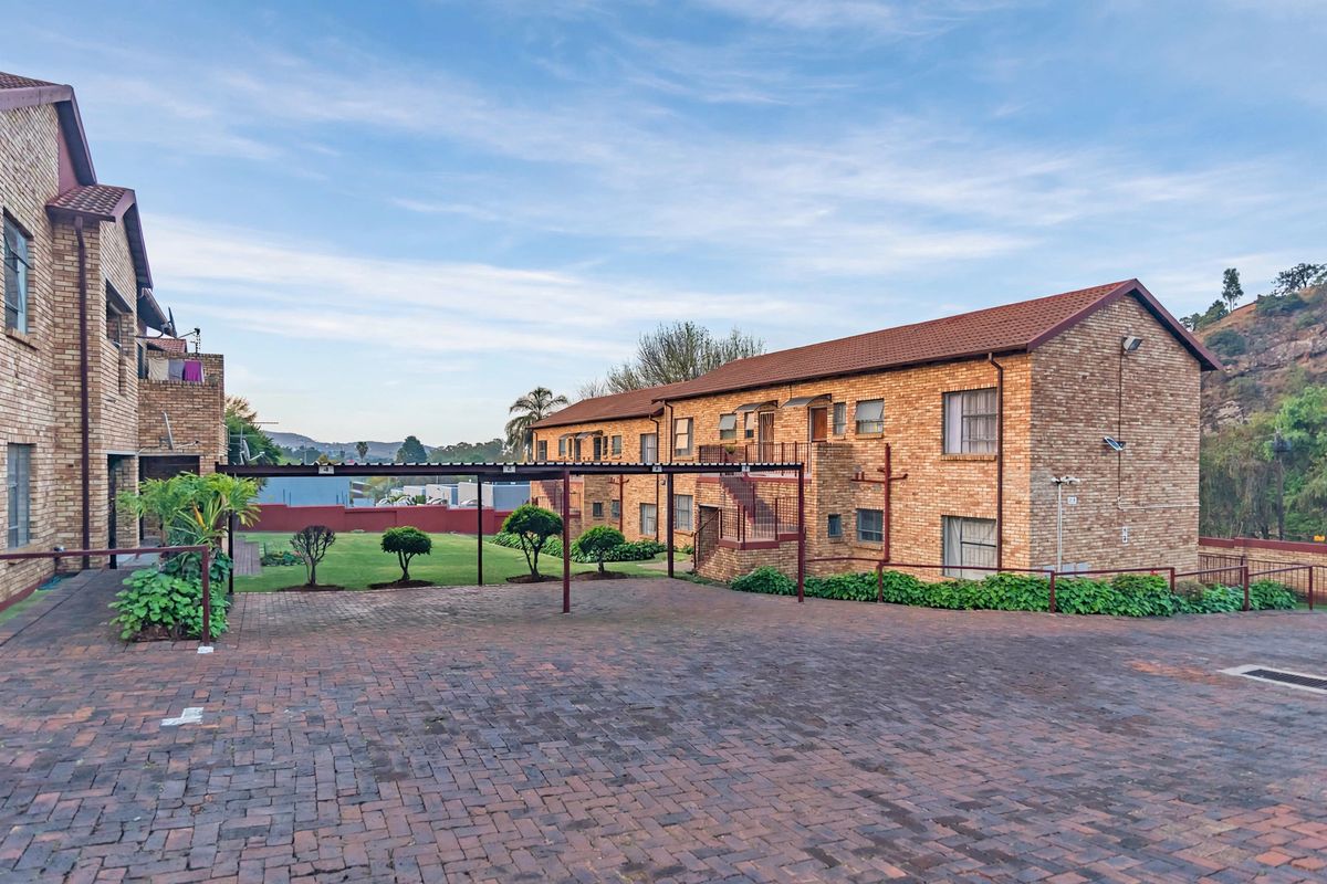 Exterior view of a face brick apartment complex featuring a paved driveway, covered carport area, and a well-maintained communal garden space.