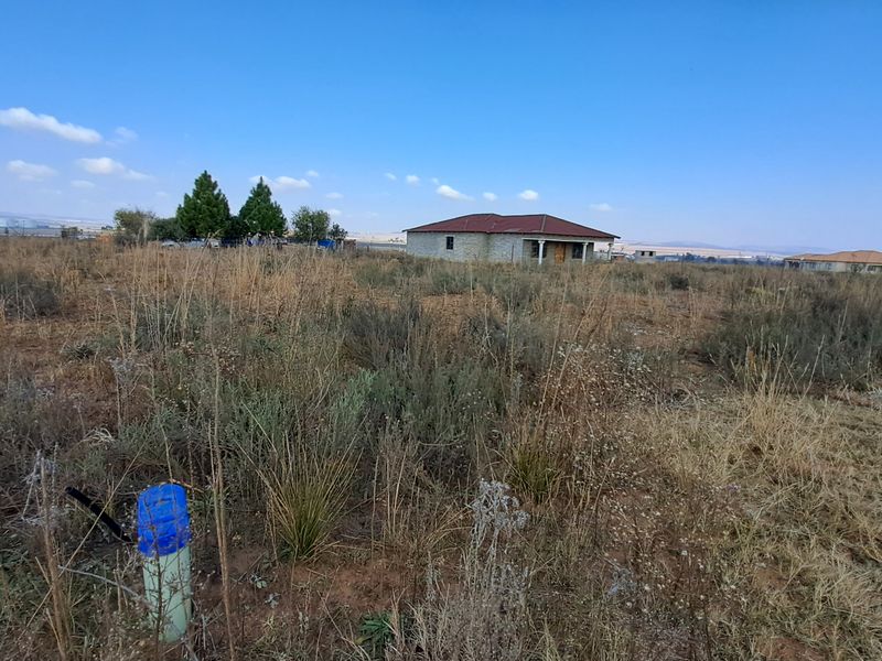 A wide-angle view of a vacant residential plot featuring natural vegetation and a visible utility pipe in the foreground.