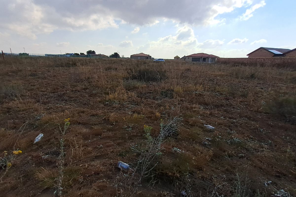A wide-angle view of a vacant residential plot featuring dry vegetation and surrounding perimeter walls in a developed neighborhood.