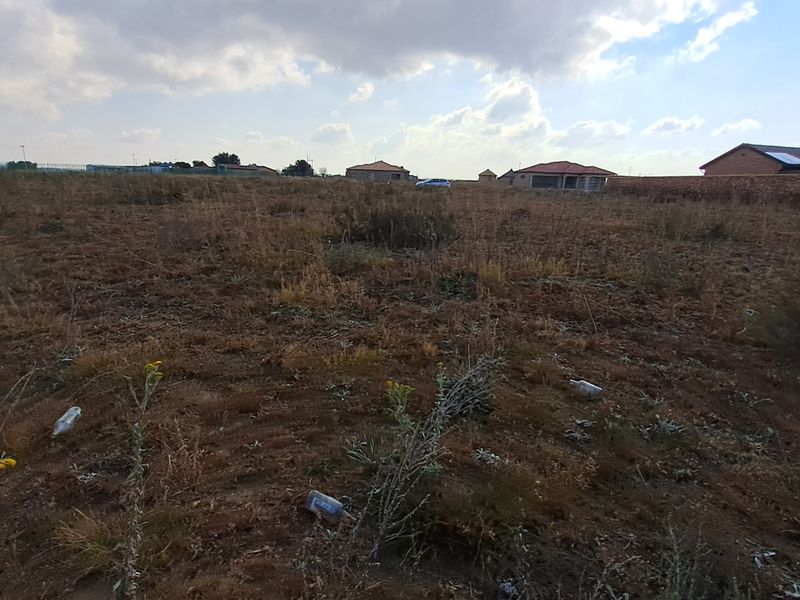 A wide-angle view of a vacant residential plot featuring dry vegetation and surrounding perimeter walls in a developed neighborhood.