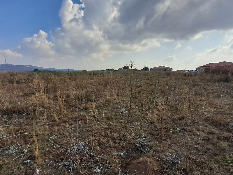 A wide-angle view of a vacant residential plot featuring natural dry vegetation and surrounding neighborhood development.