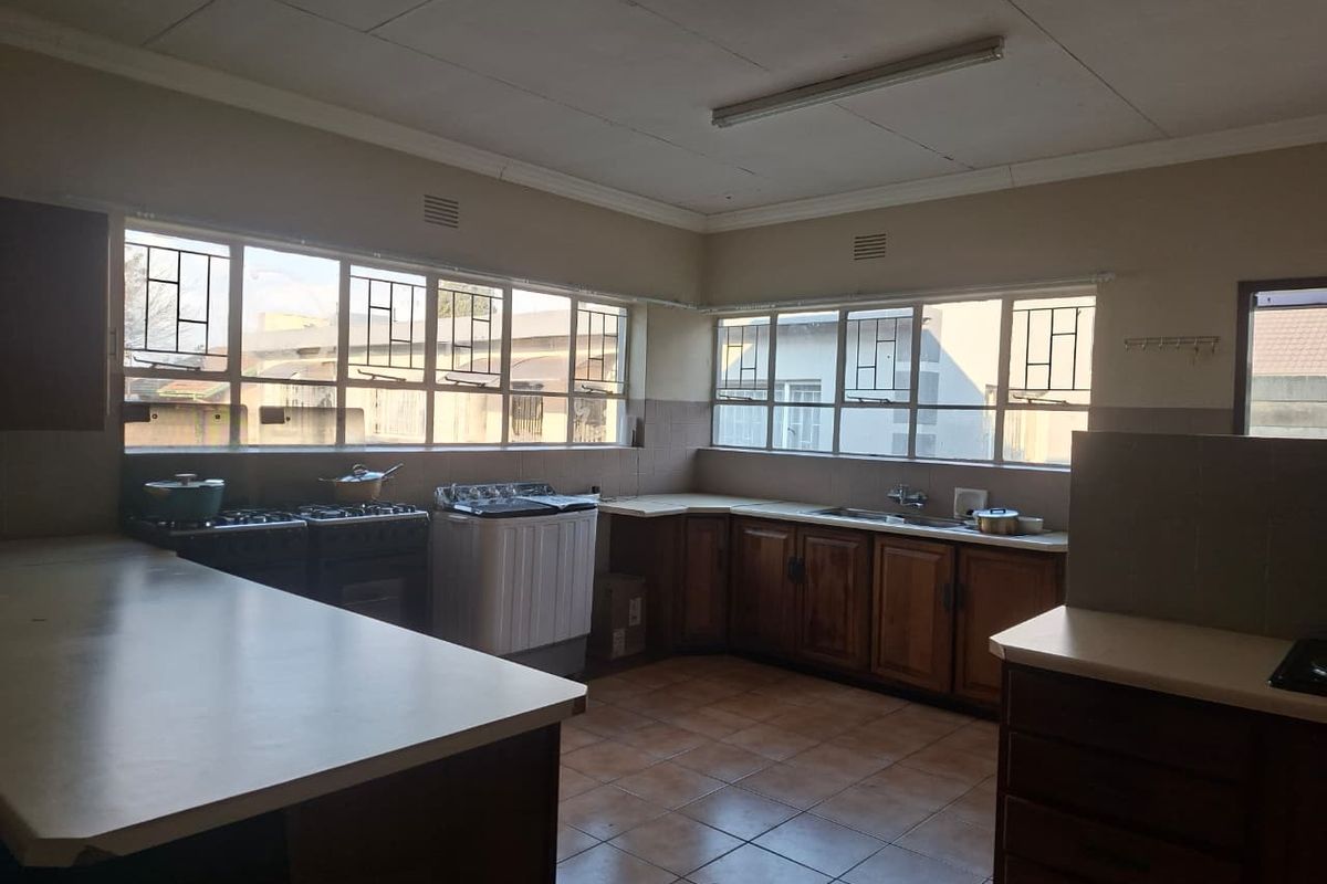 A spacious, dated kitchen featuring wooden cabinetry, laminate countertops, tiled flooring, and large windows with security bars, offering significant potential for modernization.