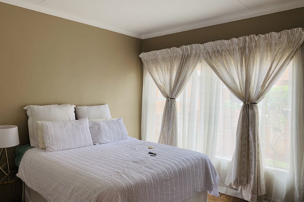 A bedroom featuring neutral wall tones, tiled flooring, and large windows dressed with layered curtains, providing natural light and a comfortable living space.