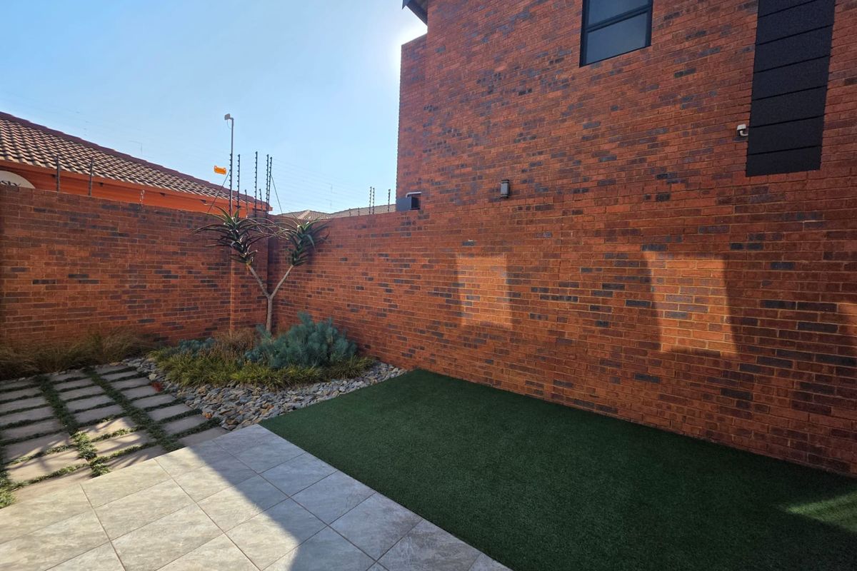 A private rear courtyard featuring a low-maintenance artificial lawn, tiled patio area, and secure face brick boundary walls with electric fencing.