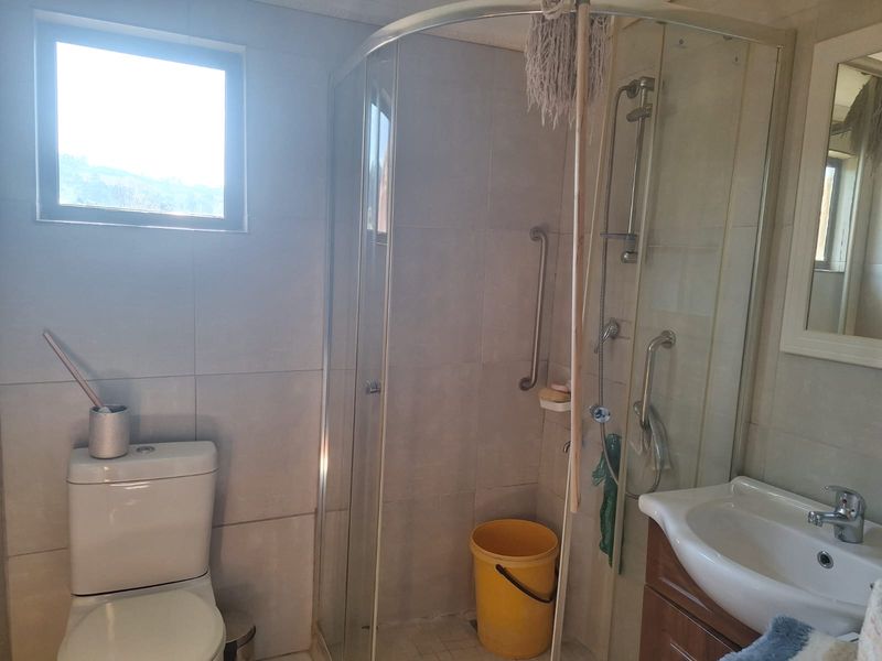 A functional bathroom featuring a corner shower enclosure, toilet, and vanity basin with tiled walls and grab rails for accessibility.