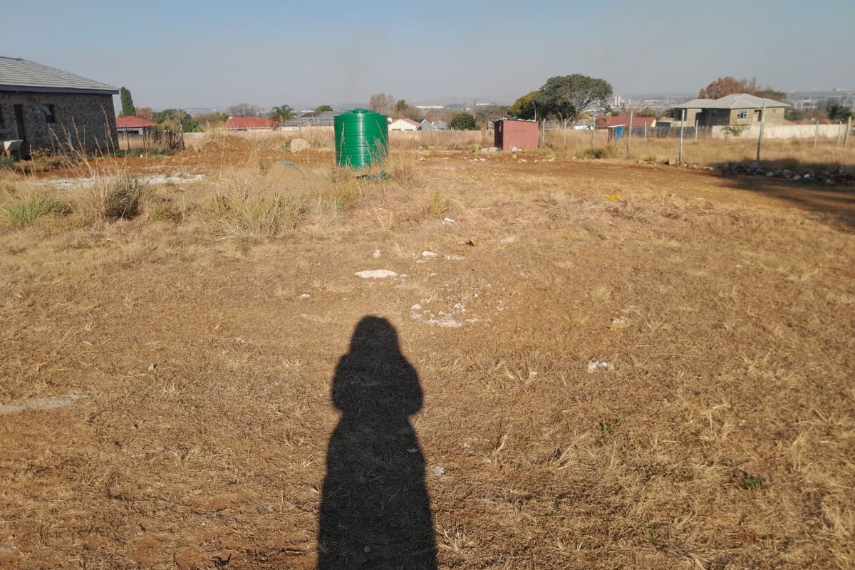A view of a vacant residential plot featuring dry grass, a water storage tank, and perimeter wire fencing.