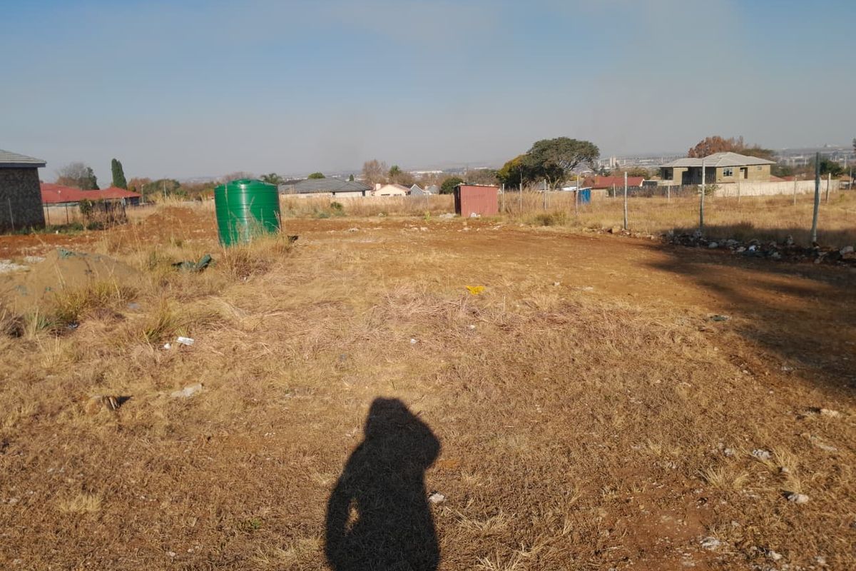 A vacant residential plot featuring dry grass, a water storage tank, and perimeter wire fencing in a suburban setting.