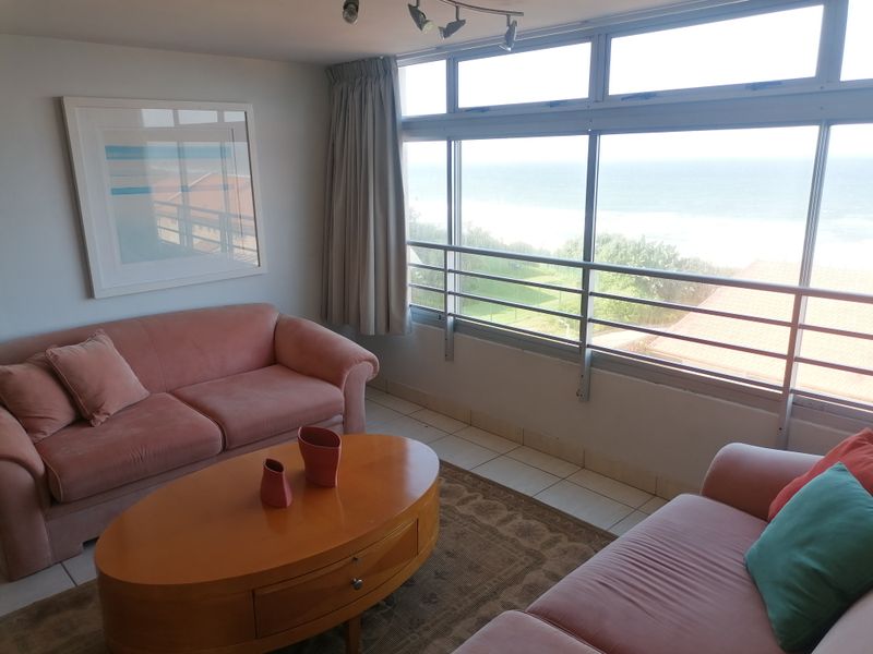 A bright living room featuring large windows with ocean views, tiled flooring, and track lighting, offering a comfortable space with natural light.