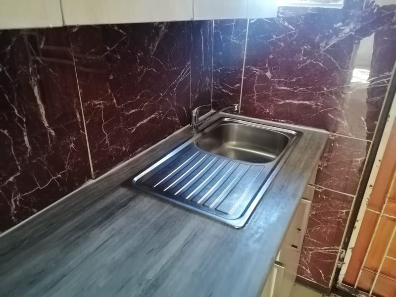 A compact kitchen area featuring a stainless steel sink, laminate countertop, and distinctive dark marble-effect wall tiling.