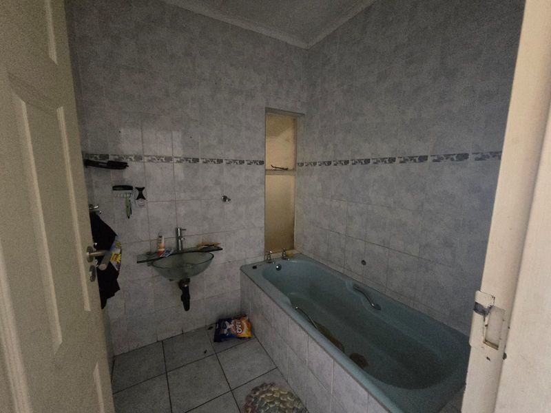 A dated bathroom featuring a built-in bathtub, wall-mounted basin, and full-height wall tiling, requiring cosmetic updates to modernize the space.