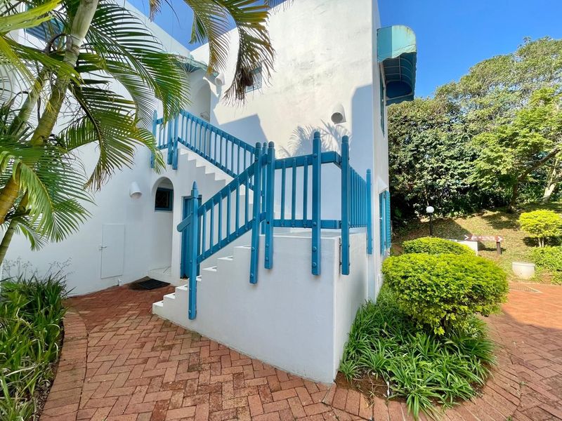 Exterior view of an apartment building entrance featuring a brick paved walkway, white facade, and a distinctive blue painted staircase and balustrade.