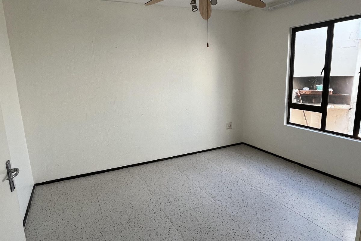 A bright, empty bedroom featuring neutral tiled flooring, a ceiling fan, and a large window providing natural light.