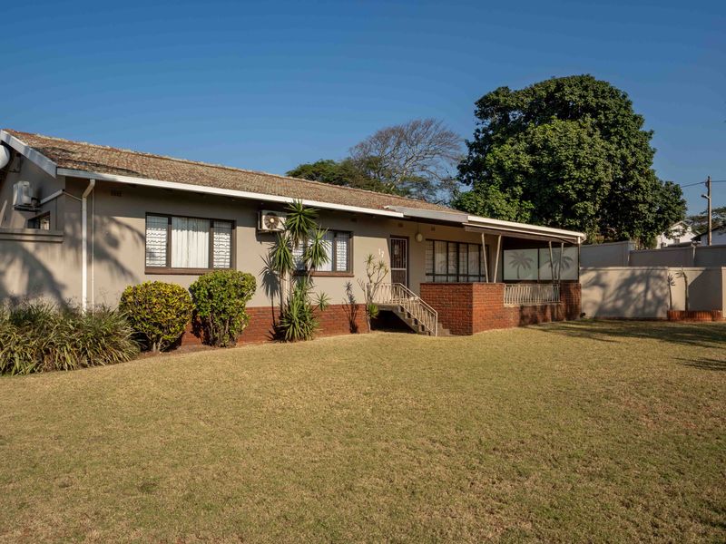 A well-maintained single-story house featuring a brick facade, tiled roof, and a covered patio area overlooking a spacious lawn.