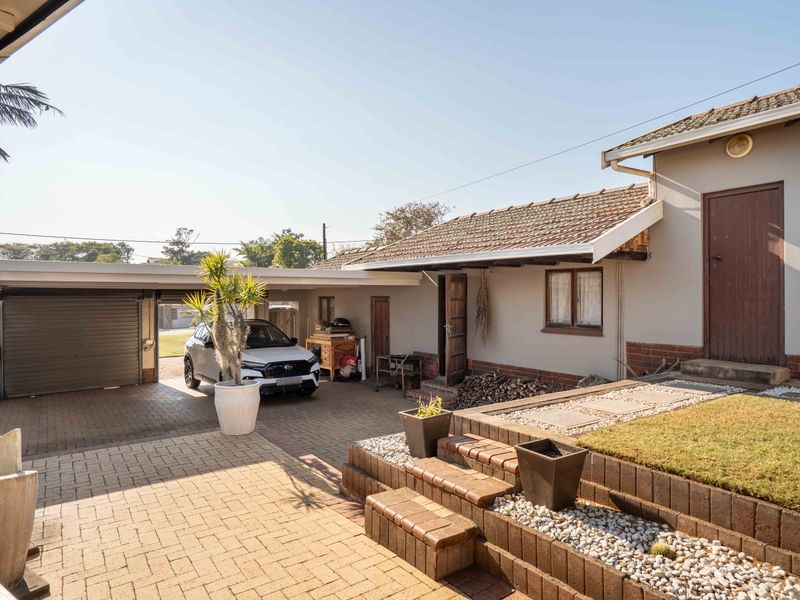 Rear exterior view featuring a paved driveway, carport, and tiered garden steps leading to the main dwelling with a tiled roof.
