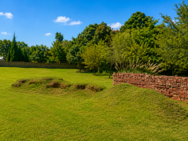 A spacious, well-maintained residential garden featuring a lush lawn, mature trees, a brick boundary wall, and a stack of loose bricks.