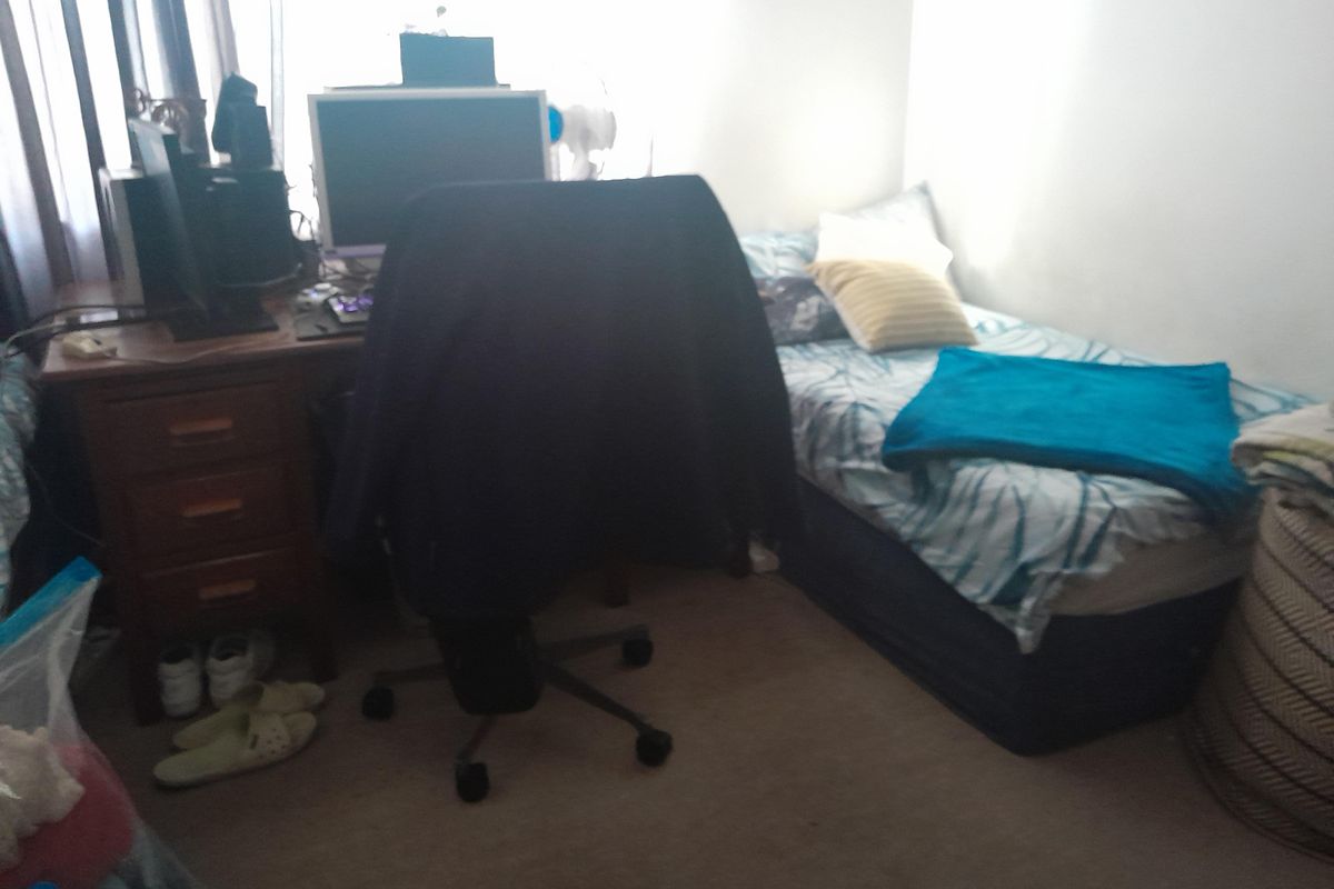 A compact bedroom featuring carpeted flooring and a window with curtains, currently utilized as a combined sleeping and workspace area.