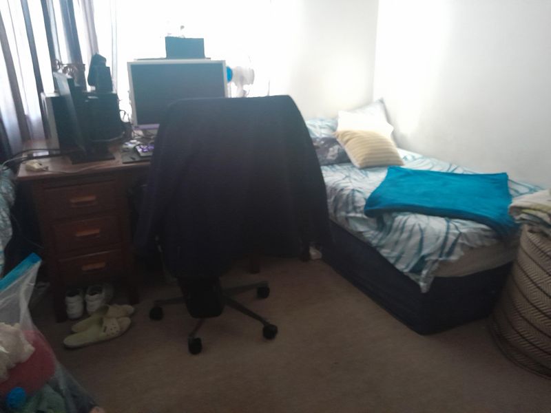 A compact bedroom featuring carpeted flooring and a window with curtains, currently utilized as a combined sleeping and workspace area.