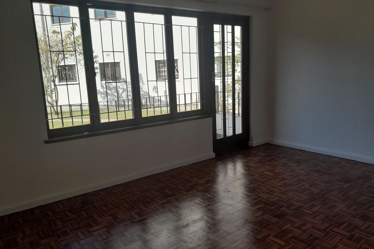 A bright, empty living room featuring classic parquet flooring, large windows with security bars, and a glass-paneled door leading to the exterior.