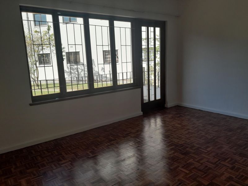 A bright, empty living room featuring classic parquet flooring, large windows with security bars, and a glass-paneled door leading to the exterior.