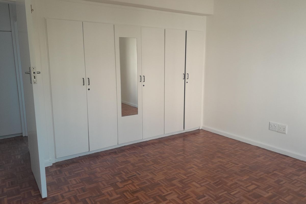 A clean, empty bedroom featuring classic parquet flooring and extensive white built-in cupboards with a central mirror panel.