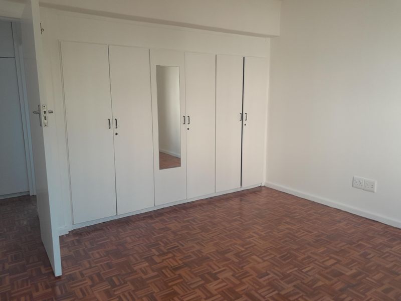 A clean, empty bedroom featuring classic parquet flooring and extensive white built-in cupboards with a central mirror panel.