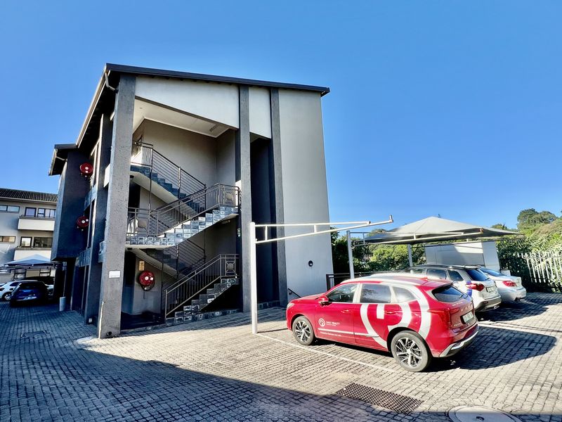 Exterior view of an apartment building featuring a paved parking area, covered carports, and an external staircase providing access to the units.