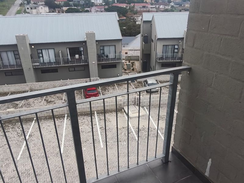 A private balcony featuring a metal balustrade, tiled flooring, and an exposed brick wall, overlooking the apartment complex parking area.
