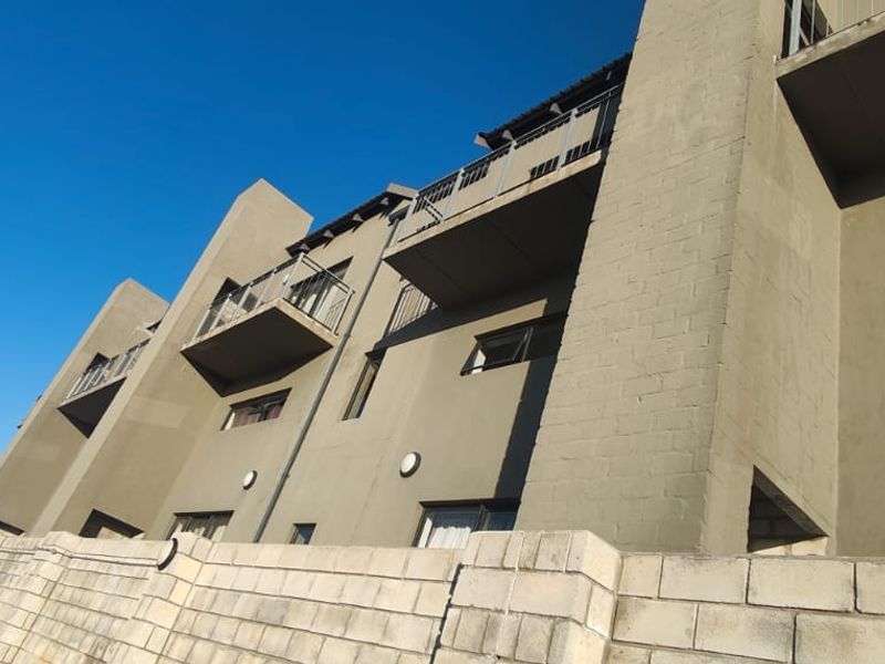 A low-angle view of the apartment building exterior, featuring multiple balconies with metal railings and a concrete block retaining wall.