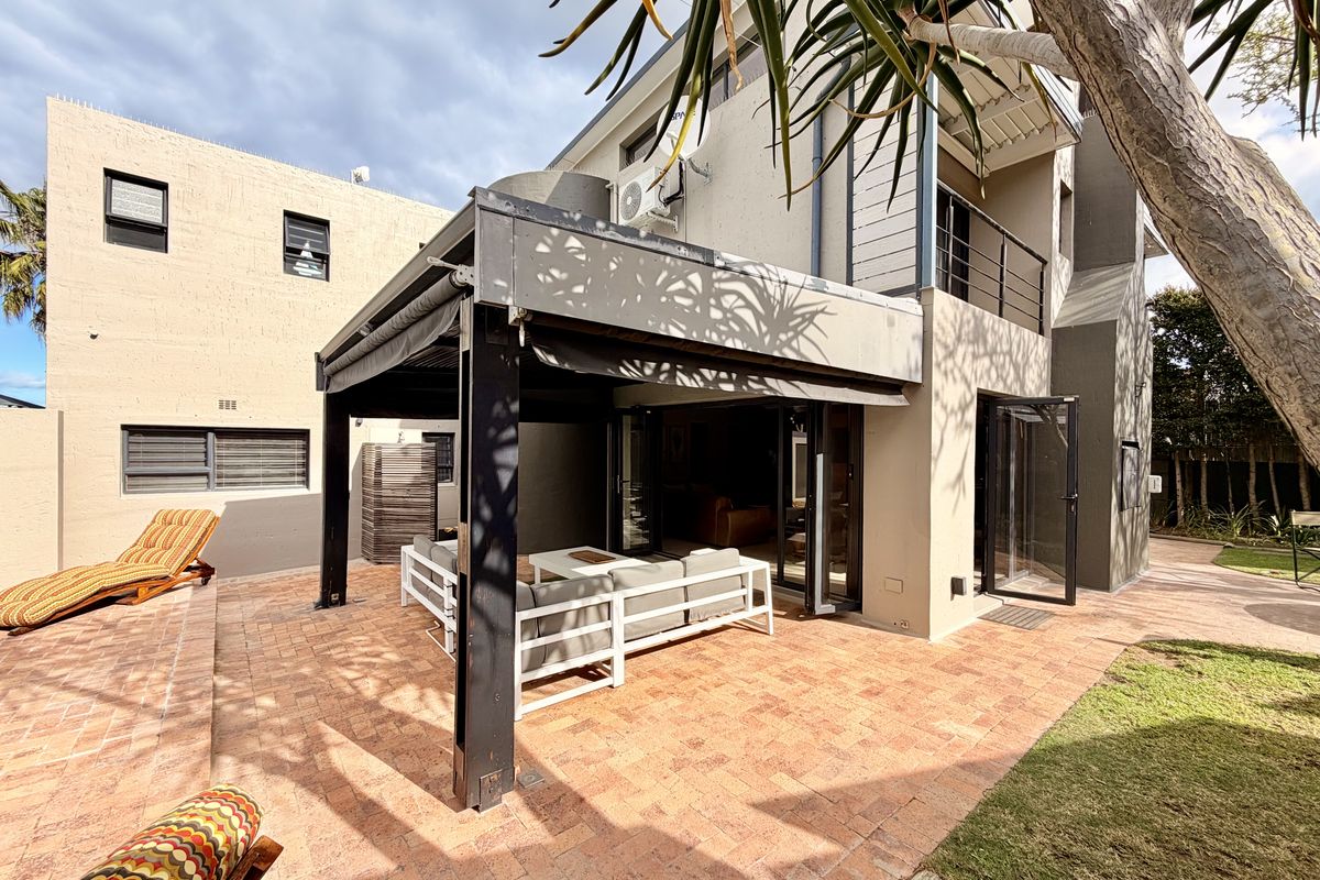 Rear exterior view featuring a paved patio area with a covered structure, balcony access, and established garden surroundings.