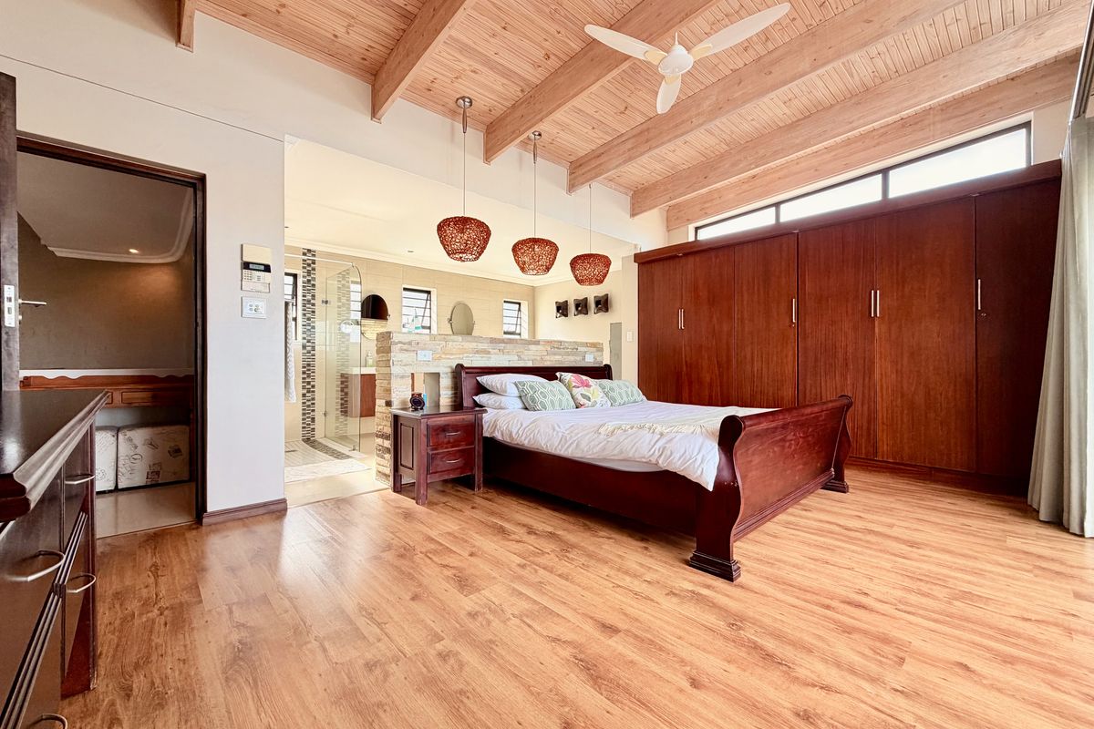 Spacious bedroom featuring exposed timber ceiling beams, laminate flooring, and extensive built-in wooden cabinetry, with an open-plan flow to the en-suite bathroom.