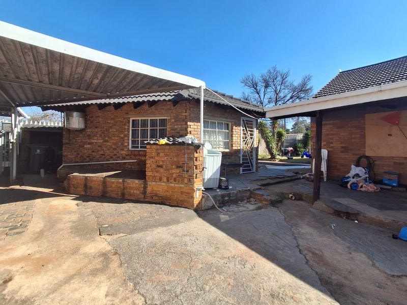 Rear exterior view of the property featuring a brick facade, covered patio area, and concrete paving, showing signs of wear and maintenance requirements.