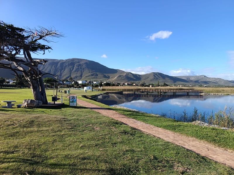 Scenic view of a tranquil lake, wooden footbridge, and surrounding mountain landscape, featuring a well-maintained lawn and brick pathway.