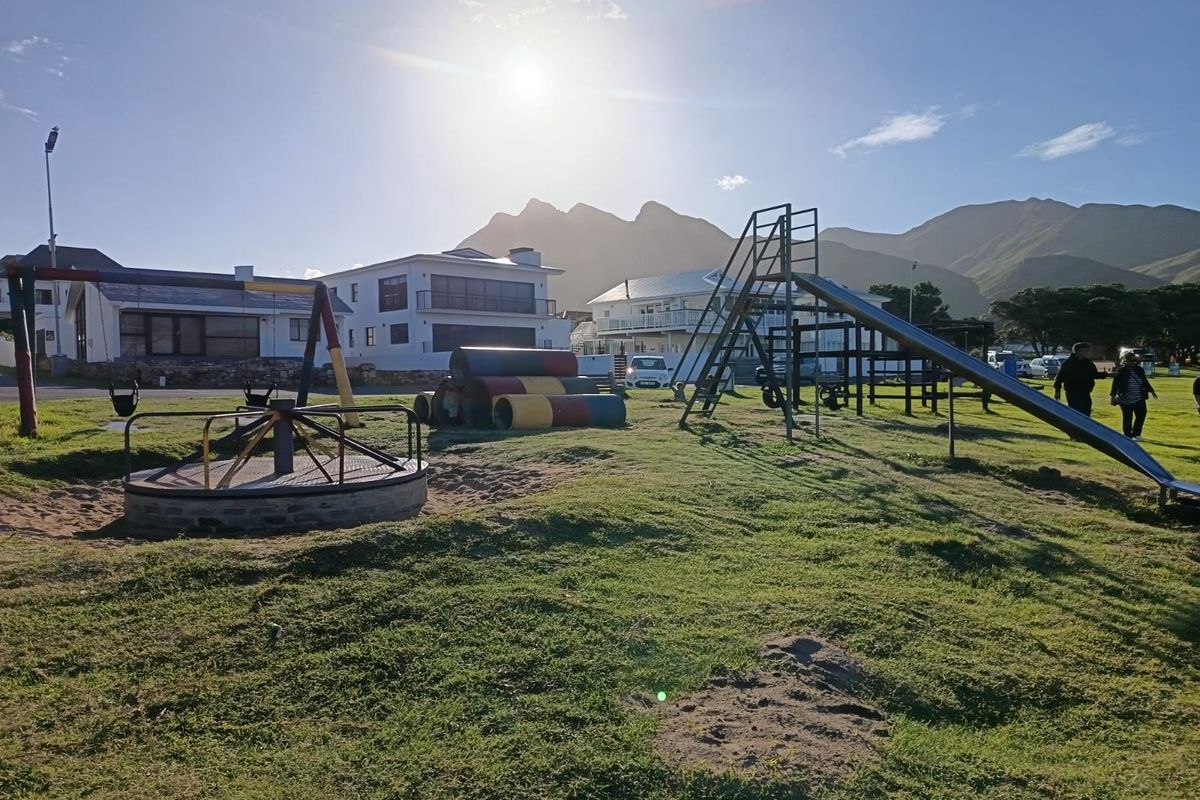 A public park area featuring playground equipment including a slide, merry-go-round, and tunnels set against a mountain backdrop.
