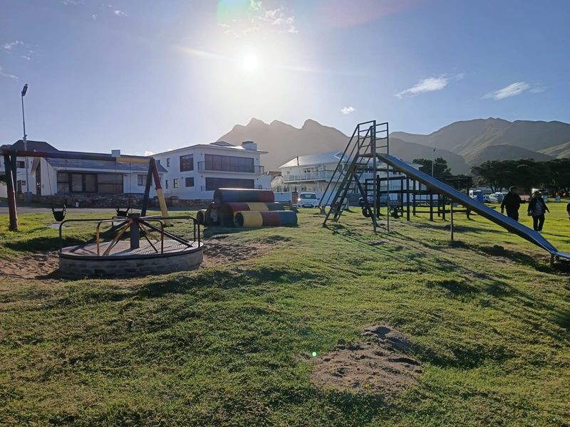 A public park area featuring playground equipment including a slide, merry-go-round, and tunnels set against a mountain backdrop.
