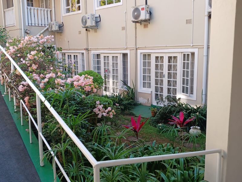 Rear exterior view of the apartment complex featuring landscaped garden beds, paved walkways with safety railings, and multiple ground-floor french door access points.