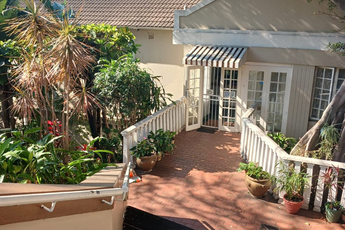 A rear exterior view featuring a brick-paved walkway leading to a building entrance with a striped awning and white railings.