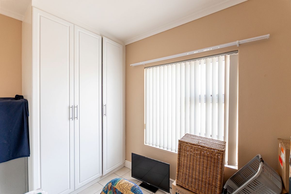 A bedroom featuring white built-in cupboards, tiled flooring, and a window fitted with vertical blinds.