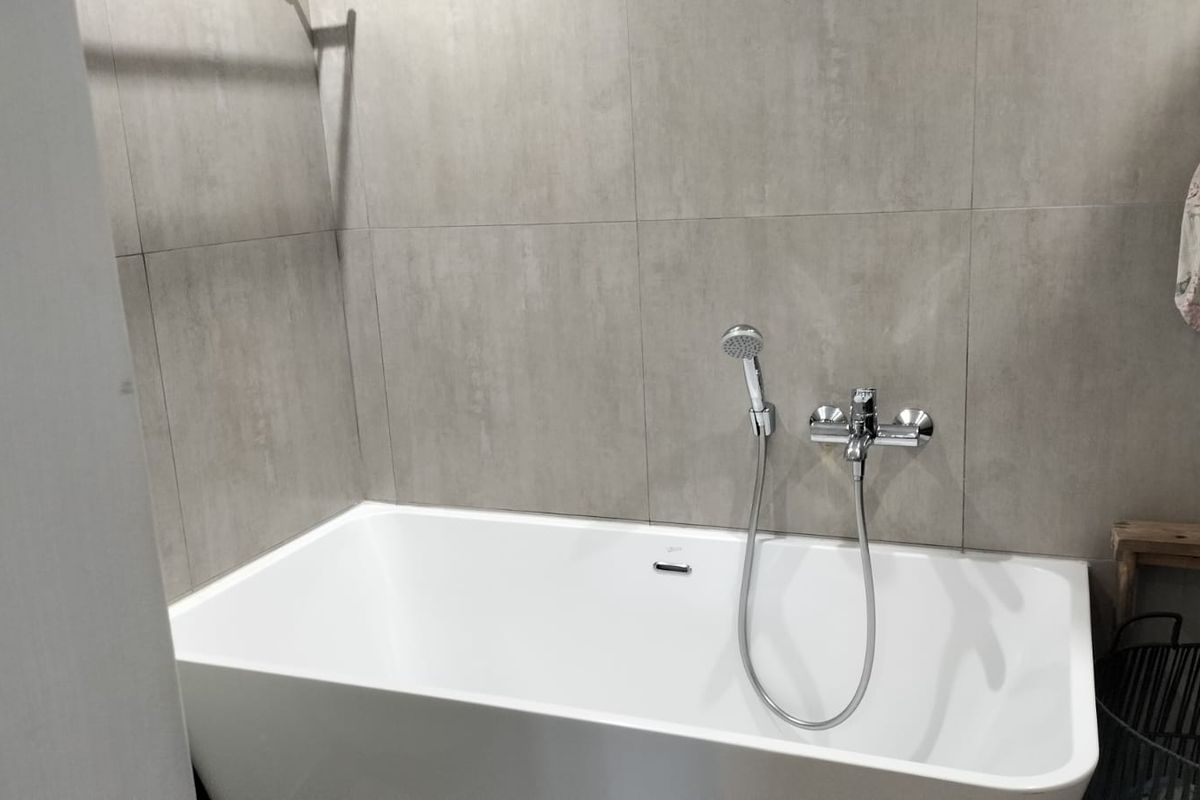 A modern bathroom featuring a clean white bathtub with a chrome shower mixer and handheld shower head against neutral grey wall tiles.
