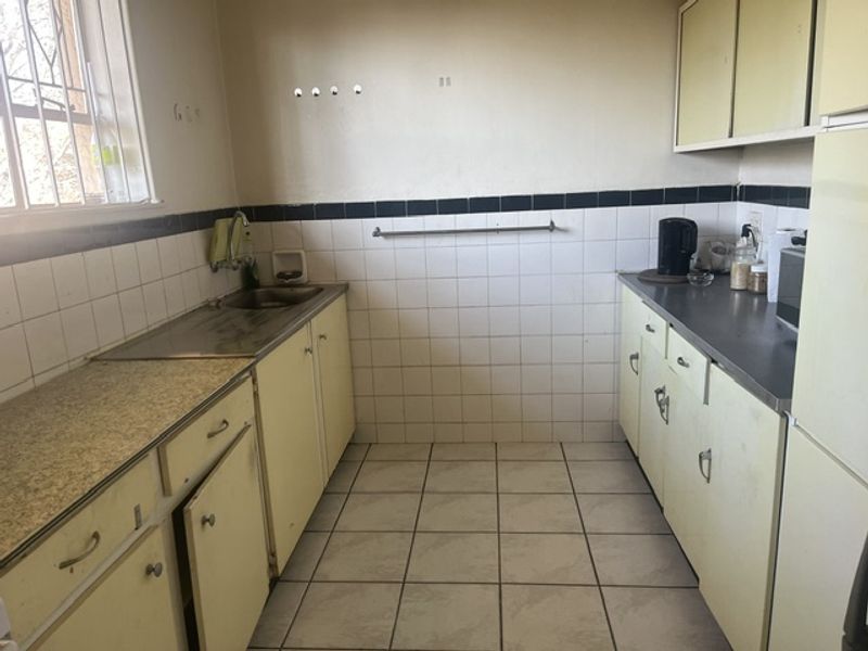 A dated kitchen featuring original cabinetry, laminate countertops, ceramic wall and floor tiles, and a stainless steel sink with window security bars.