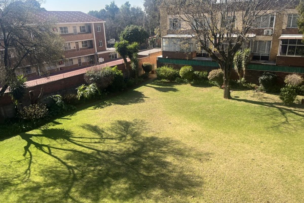 An elevated view of a communal rear garden area featuring a well-maintained lawn, mature trees, and brick boundary walls within an apartment complex.