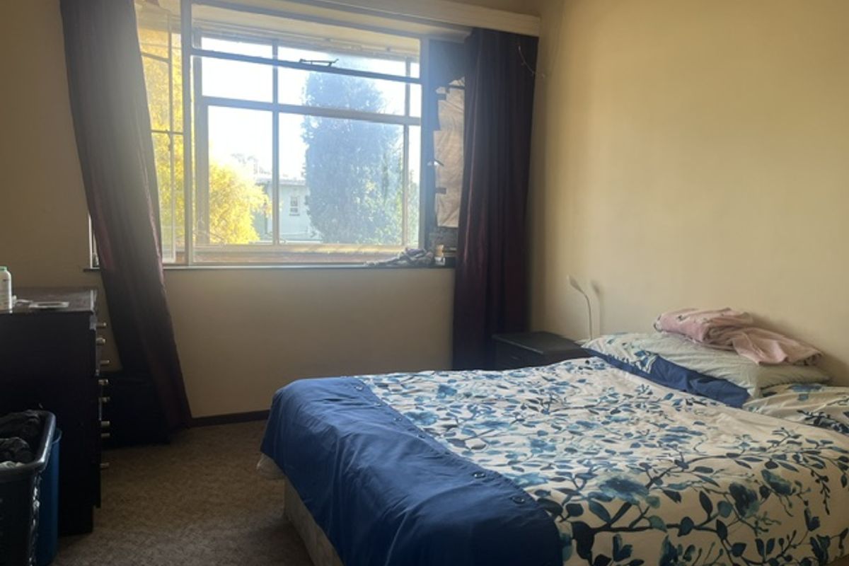 A bedroom featuring carpet flooring and a large window with natural light, currently requiring cosmetic updates to the walls and window fixtures.