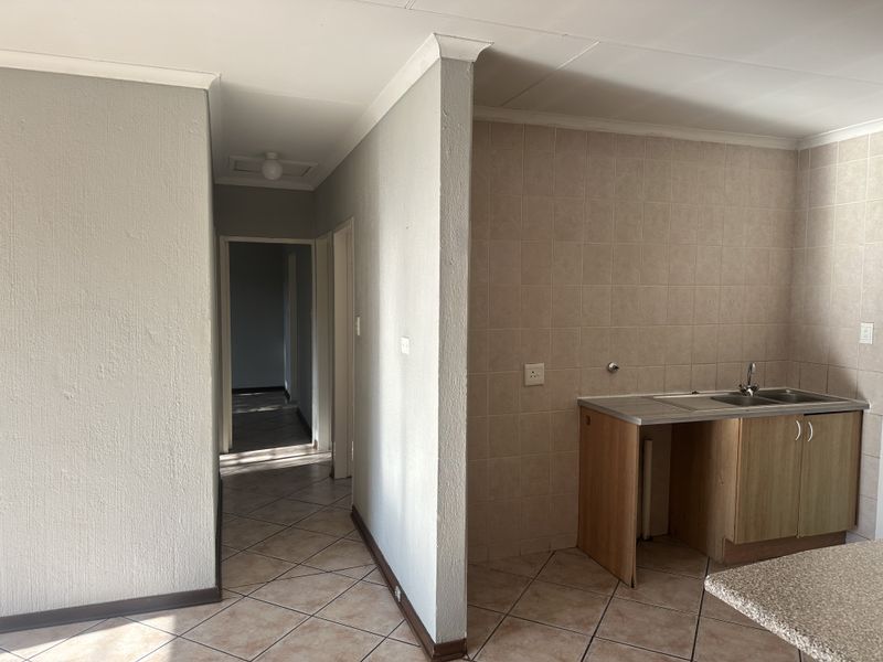 A compact kitchen area featuring tiled splashbacks, a stainless steel sink, and base cabinetry, leading into a hallway with tiled flooring.
