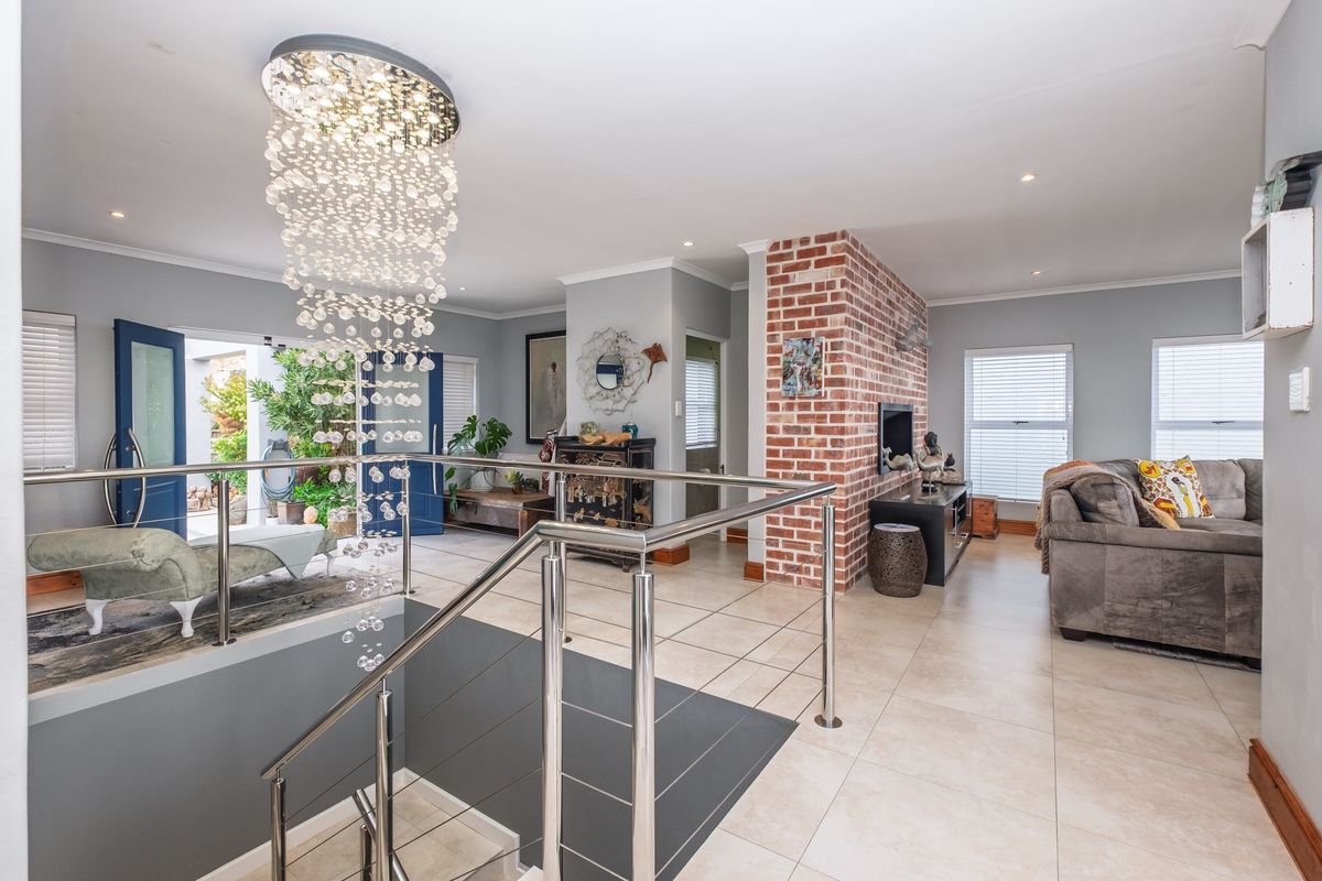 Spacious living area featuring an exposed brick wall, modern stainless steel balustrade, and a striking crystal chandelier, with large windows providing natural light.