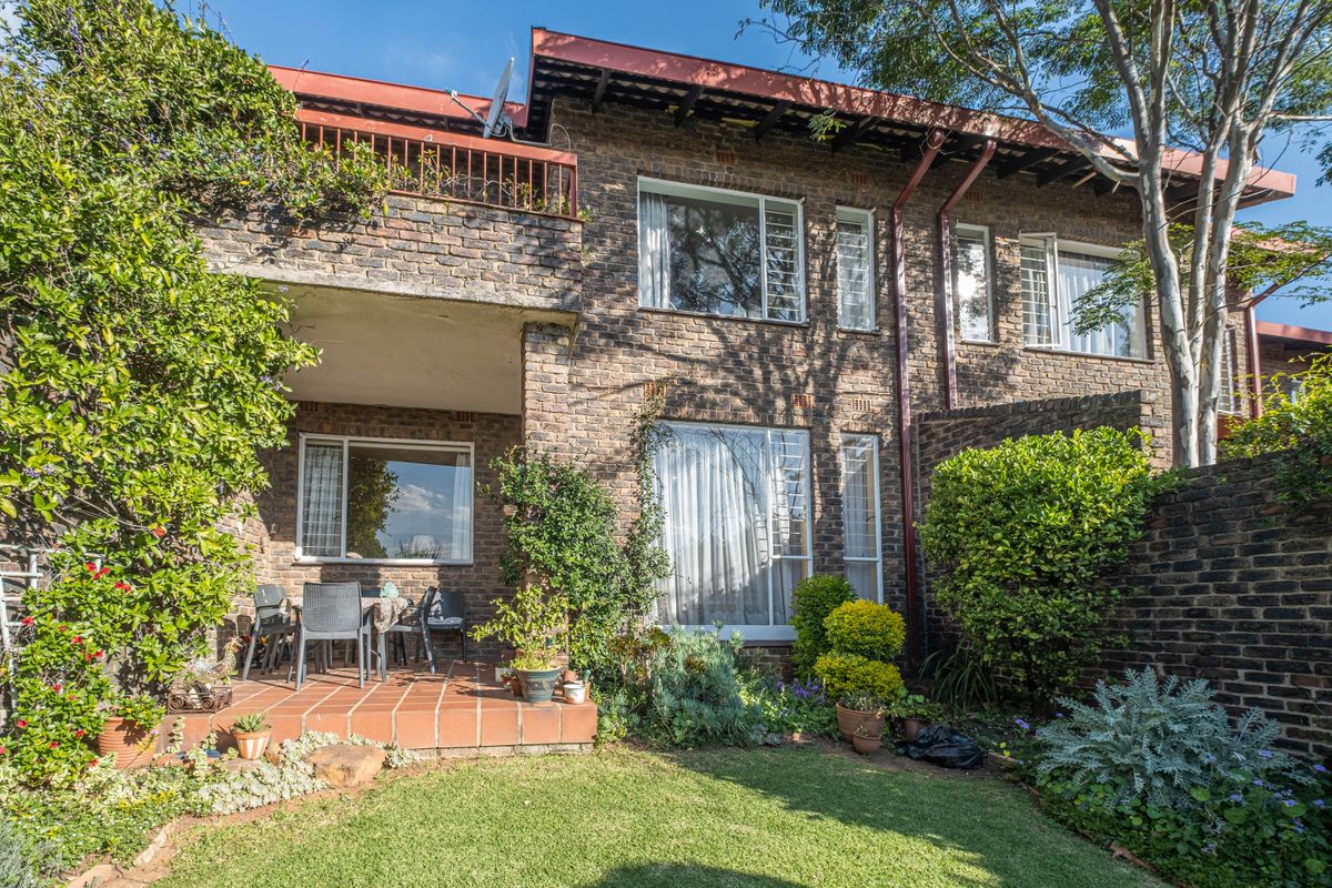 Rear view of a double-story townhouse featuring a face brick facade, a covered patio area, an upper-level balcony, and a well-maintained private garden.